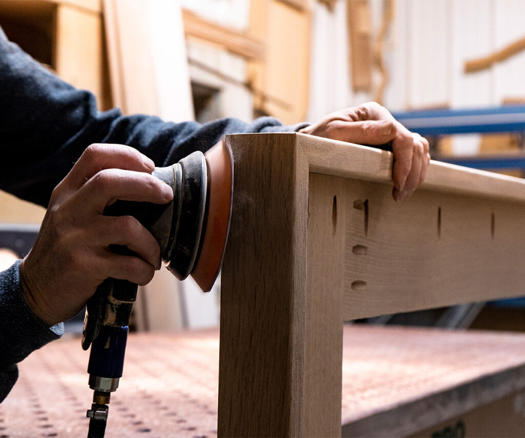 Woodworker sands solid wood table corner with a pneumatic sander.