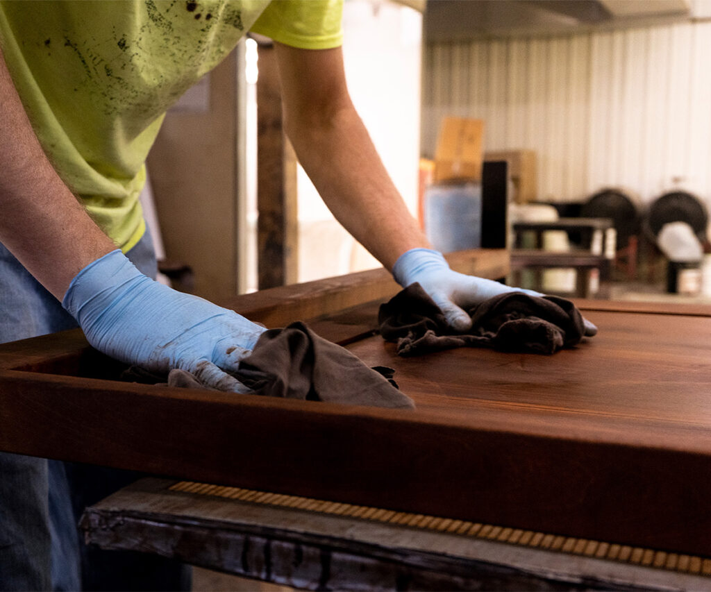 Worker hand-applies stain to solid wood tabletop with cloth.