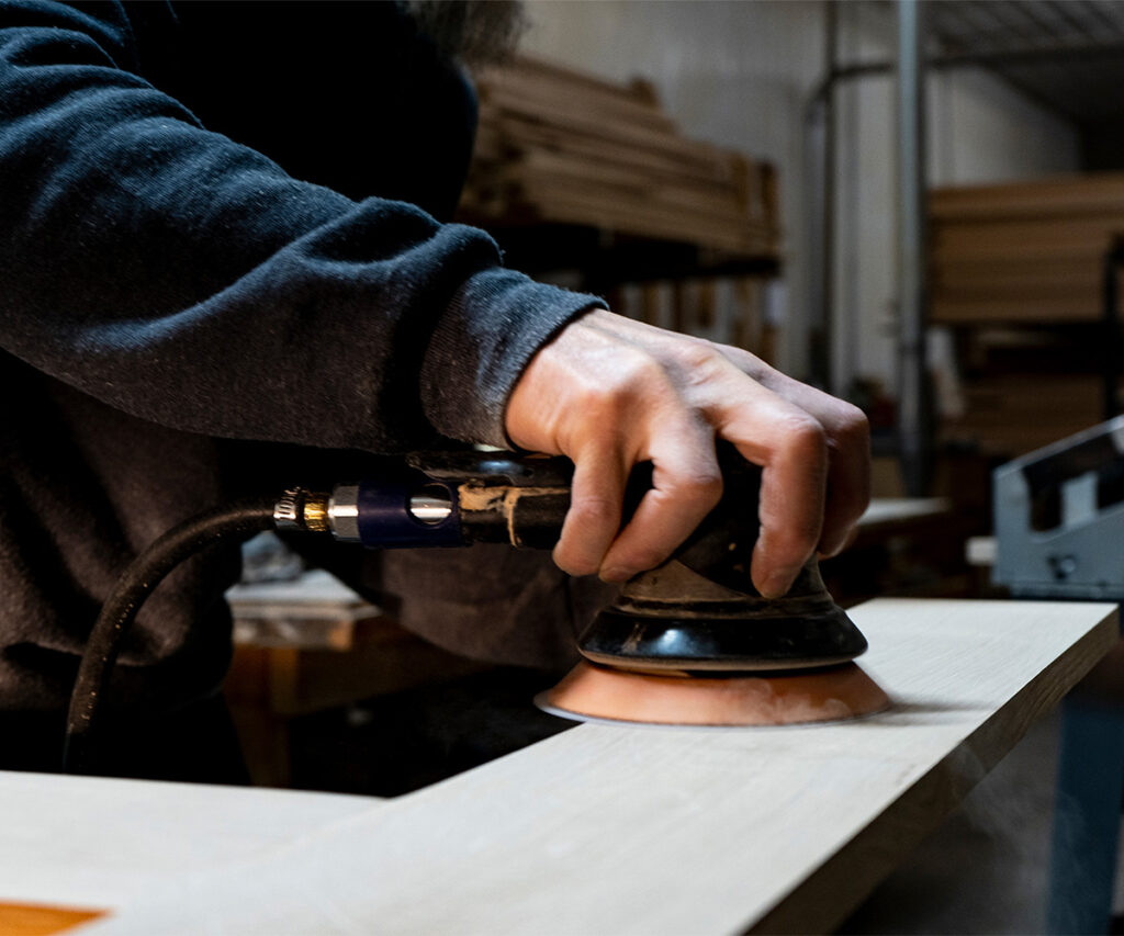 How Amish furniture is made: craftsman sands solid wood panel with orbital sander.